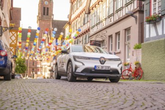 A Renault stands on a colourful street with half-timbered houses and balloons, Deer E, car sharing,