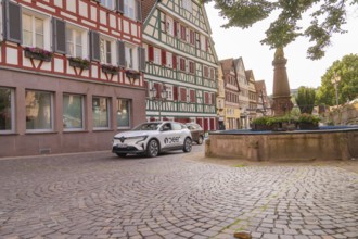 A car next to a fountain in a cobbled old town street with half-timbered facades and green plants,