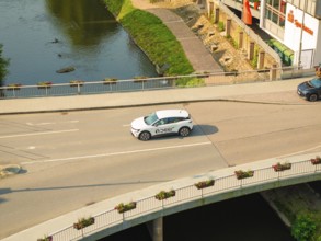 A white car drives on a bridge over a river, surrounded by urban architecture and nature, Deer E,