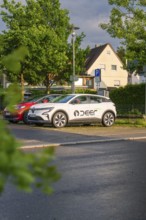 A white electric car parked in a car park in front of a house in a green residential area, Deer E,
