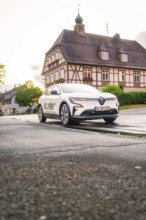 A white electric car parked in front of a traditional building with a bell tower in an old town,