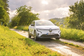 A white electric car drives along a rural gravel road surrounded by trees in sunlight, Deer E, car