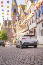 A car drives through a colourfully decorated old town with a church in the background, Deer E, car