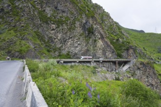 A truck crosses a bridge along steep rock faces in a green mountain landscape, damaged concrete