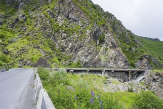A road runs along steep rock faces, with a bridge and green vegetation, damaged concrete road