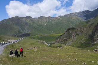 Three people sit in a meadow in front of a mountain range under a cloudy sky, left truck queue on