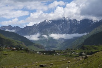 Impressive snow-capped mountains rise above a green valley with clouds in the sky, Kazbegi,
