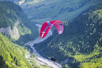 A paraglider floats over a narrow, green valley region with a fast-flowing river, paraglider near