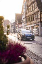 Black car in quiet old town with half-timbered houses and warm sunlight, MG4 electric car, Deer