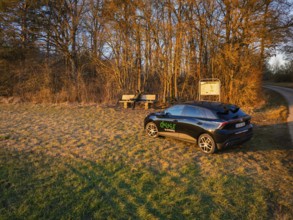 Car parked in rural landscape with benches at sunrise, surrounded by trees, MG4 electric car, deer