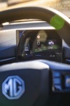 Vehicle display next to steering wheel with brand logo, showing speed and charging information, MG4