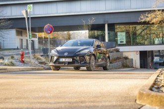 Black car drives in front of a modern building backdrop during the day, road signs show the way,
