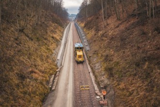 Yellow train and workers on a long railway line through autumnal forest, using a tamping machine at