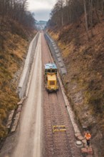 Yellow train on rails with workers in street surrounded by bare forest, using tamping machine at