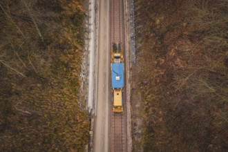 Aerial view of a yellow train traveling on rails surrounded by bare trees, using a tamping machine