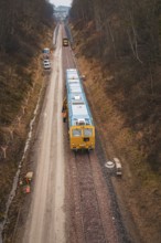 Yellow train on a construction site in a wintry landscape, surrounded by trees, using a tamping