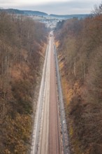 A long railway line leads through a hilly autumn landscape with bare trees, using a tamping machine