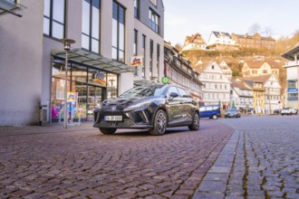 Close-up of a black car on an urban street with modern buildings in the background, MG4 electric