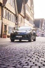 Black car drives on a cobblestone street lined with half-timbered houses and urban architecture,
