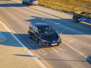 A car is parked on a wide street, illuminated by the evening sun, surrounded by open fields, MG4
