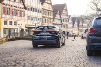 Car drives through a paved street surrounded by historic half-timbered houses, MG4 electric car,