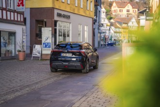 Car driving through a narrow city street surrounded by colorful buildings in the background, MG4