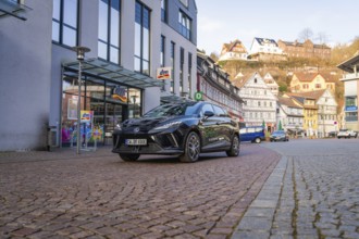 Black car in a modern city with paved streets and traditional houses in the background, MG4