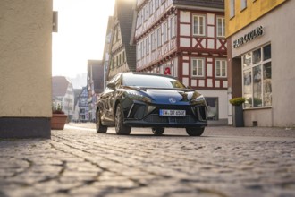Black car drives through a paved street in a historic town with half-timbered houses, illuminated