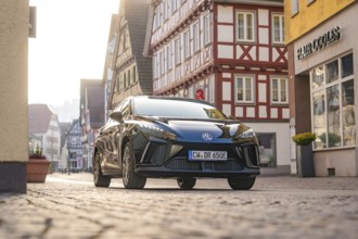 Black car on a paved street in a historic town with half-timbered houses in sunny weather, MG4