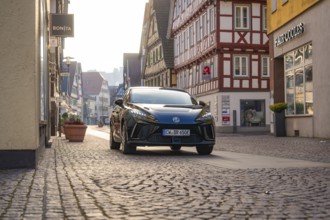 Black car drives through a paved street in a historic city environment with half-timbered houses,