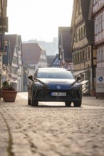 Black car on an empty, paved road surrounded by traditional building facades in sunshine, MG4