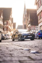 A car drives through a picturesque old town with a church in the background in sunlight, MG4