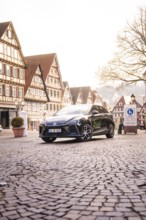 A car is parked in a paved old town and is surrounded by historic buildings and lanterns, MG4