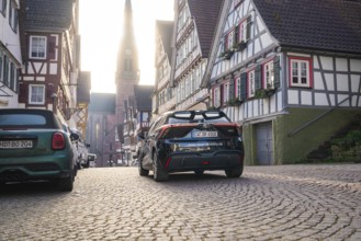 Timbered road with parked cars and church tower in the background in daylight, MG4 electric car,