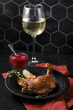 Duck confit with roasted pears on a black plate next to fruit jam and a glass of white wine. The