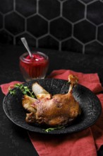 Roasted duck leg with crisp skin and tender meat on a stone plate. A small jar filled with a