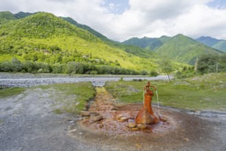 A rusty spring flows onto a gravel riverbed surrounded by green mountains, mineral spring near