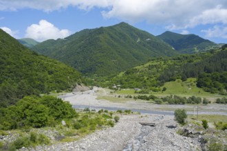 A riverbed leads through green mountains under a partly cloudy, blue sky, landscape near Chartali,