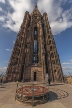 Visitors stand on a platform around the clock and admire Strasbourg Cathedral. The sky is blue with