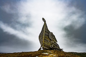 Dramatic view of an erected wooden Viking ship against a cloudy sky, sculpture by Drakkar Vardø.