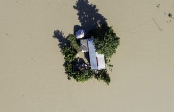 A top-down aerial view of a lone house and trees surrounded by floodwaters, capturing isolation,