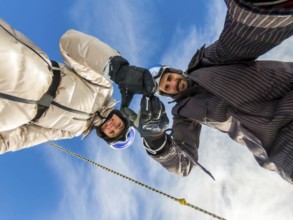 Happy couple wearing skiing gear and helmets, extending their hands upwards, forming an arch