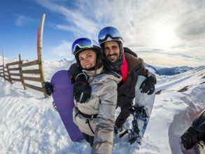 Couple smiling into camera while posing with snowboards on sunny snowy mountain slope, joyful