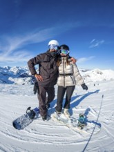 Snowboarders on a sunny mountain peak, smiling couple in helmets and goggles enjoying winter