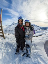 Joyful couple embracing on a snowy mountain peak, wearing helmets and goggles, holding a snowboard