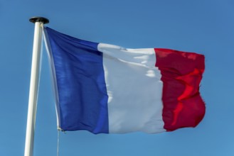 A French flag flutters in the wind on a white flagpole. The background is a clear, azure sky