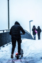 Winter weather, blowing snow, man with e-scooter trying to make his way uphill on a snowy sidewalk,