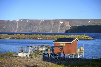 Small wooden cabin on a calm blue sea with mountain backdrop, red house on the shore with a view of