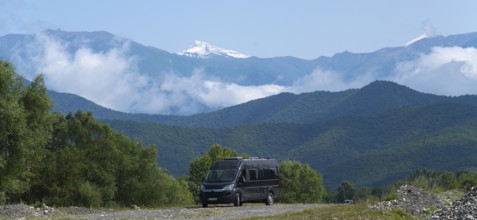 A black van on a road in a mountain valley with snowy peaks and clouds, camper in front of the
