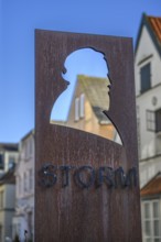 Metal sculpture with head profile of poet Theodor Storm against a city backdrop under a blue sky,